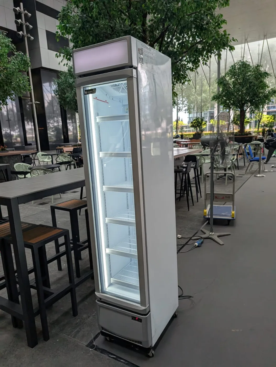 AANRS drink chiller rental Singapore, slim glass-door chiller standing on a sheltered plaza beside communal tables and stools